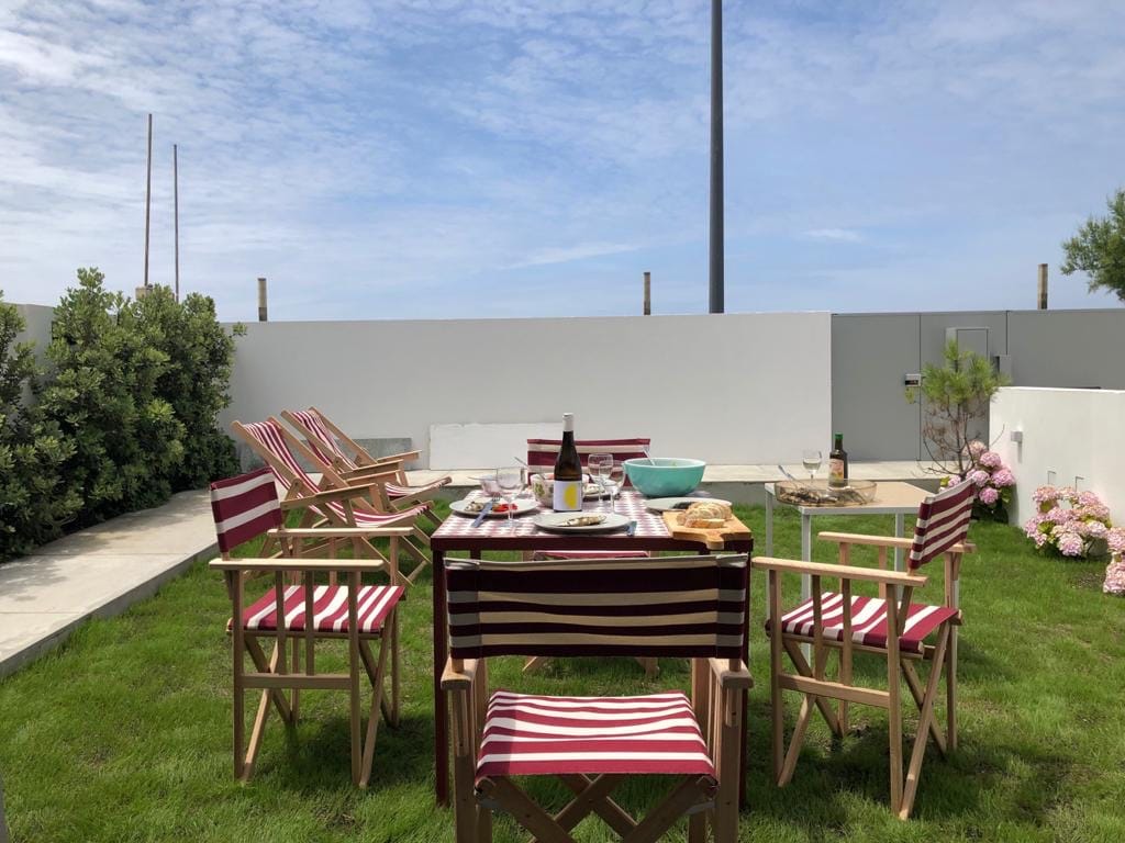 An outdoor dining area is set on the green lawn, featuring a wooden table surrounded by striped chairs. The table is adorned with plates and glasses, with a bottle of wine visible. A backdrop of low walls and foliage offers a sense of privacy.