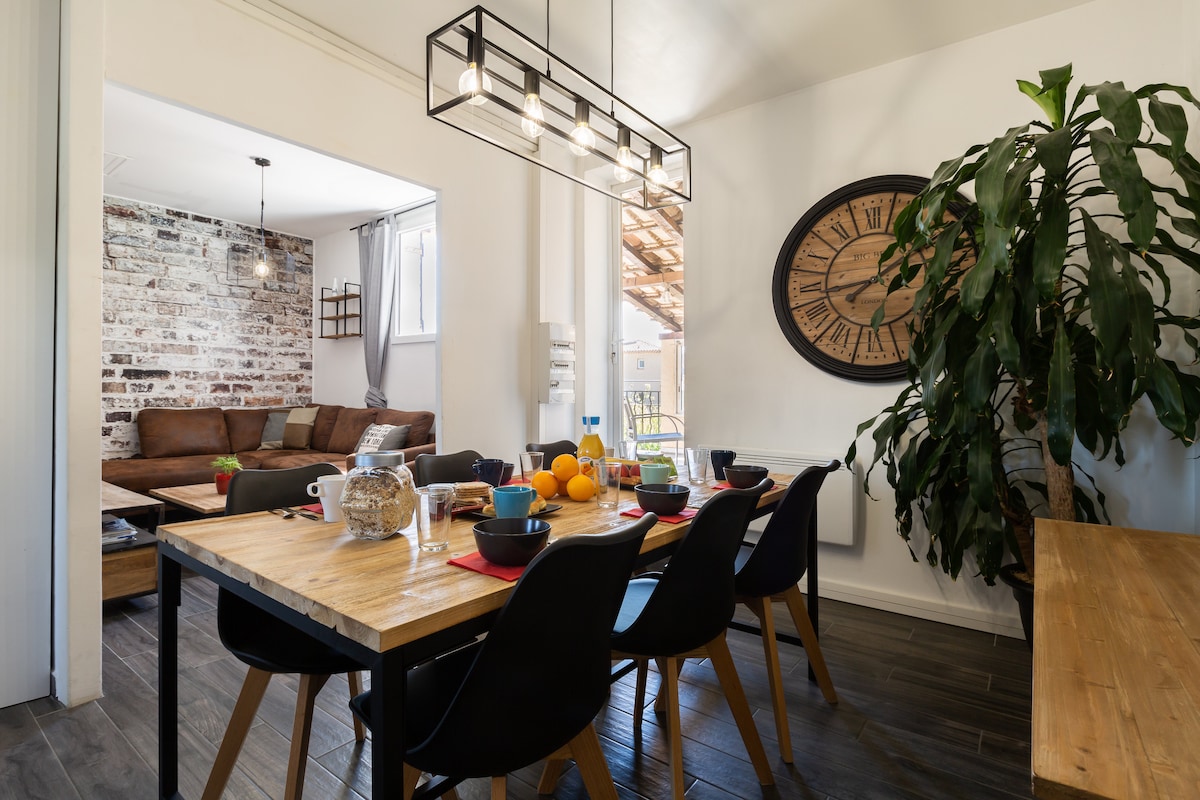 A dining area is presented with a wooden table surrounded by black chairs, set for a meal. A large clock adorns the wall, and a vibrant potted plant adds a touch of greenery. A cozy living space with a brown sofa is visible in the background.