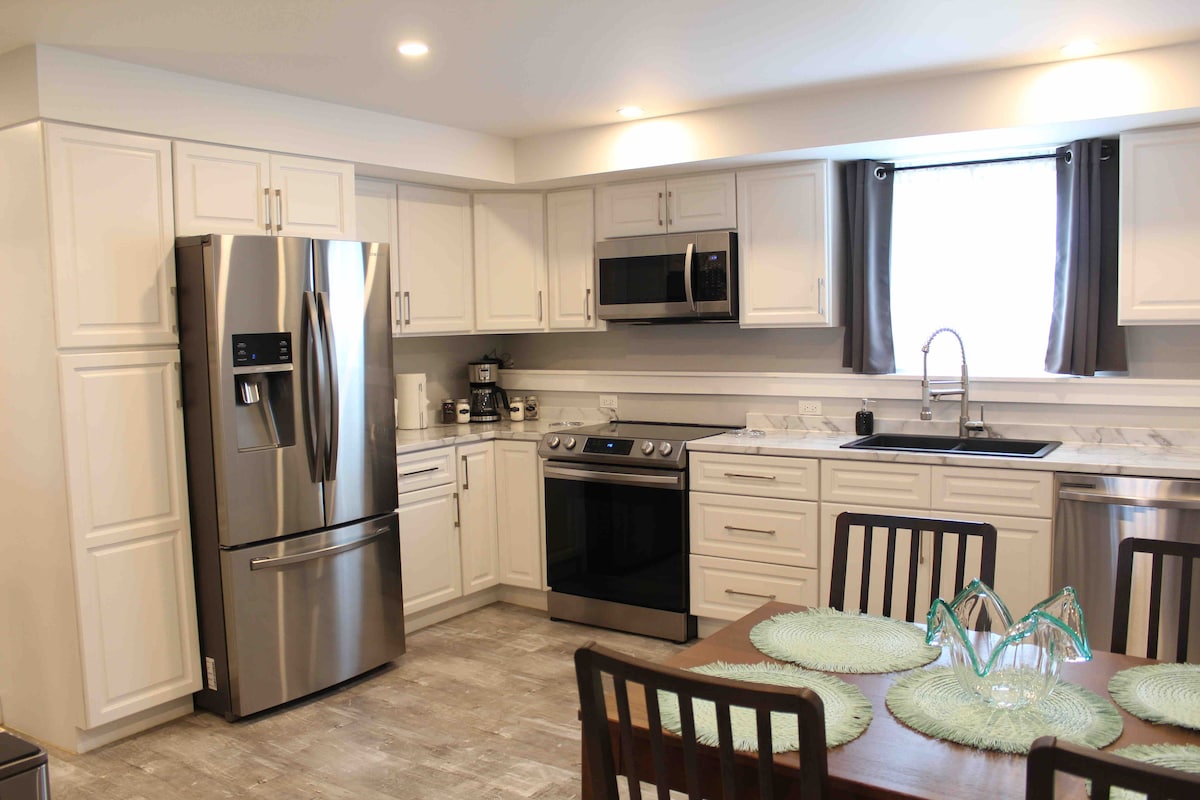 The kitchen features modern cabinetry and stainless steel appliances, including a refrigerator and oven. A dining table is set with placemats, surrounded by wooden chairs. Natural light enters through a window adorned with dark curtains.