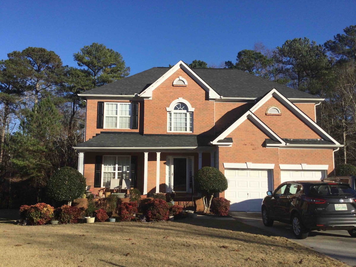 A spacious two-story brick house is set within a tree-lined yard. The home features large windows with decorative shutters and a welcoming front porch. Two garages are visible, and neatly landscaped bushes add color to the entrance against a clear blue sky.