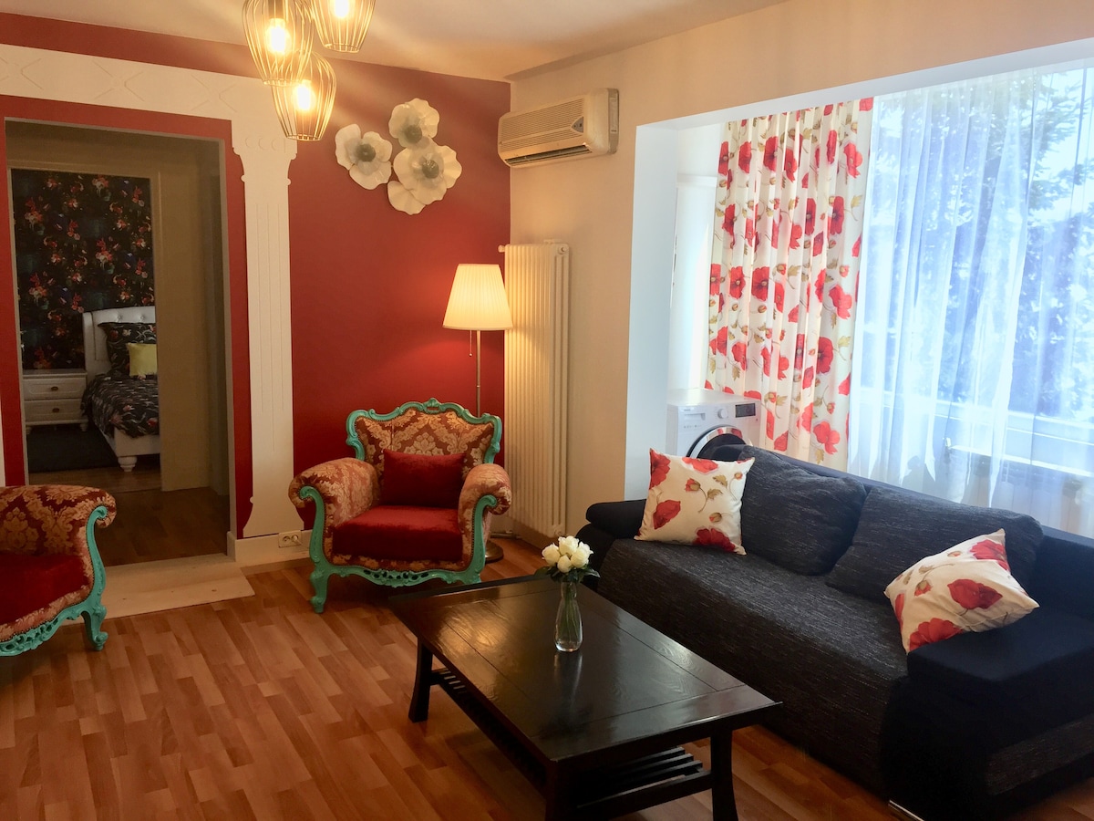 The living area features a comfortable dark sofa and a matching coffee table, with floral accents on decorative cushions. Two vintage-style chairs in bright colors are positioned near a window, allowing natural light to brighten the space. A wall adorned with floral wallpaper adds charm.