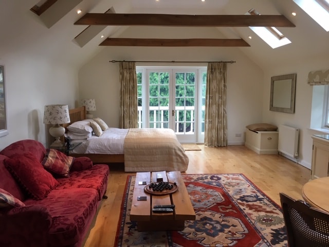 The bright and airy studio features vaulted ceilings with wooden beams. A king-sized bed is positioned beside large doors that open onto a small balcony. A cozy red sofa and coffee table are arranged in the sitting area. A decorative rug adds warmth to the wooden floor.