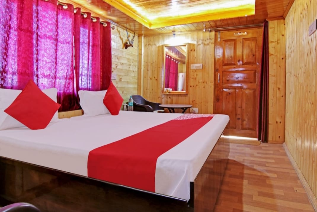 A bright guest room features wooden paneling and warm lighting. A spacious bed with a red runner is flanked by two red-cushioned pillows. Sheer curtains in rich red allow natural light to filter in. A simple mirror and chair are visible across from the bed.