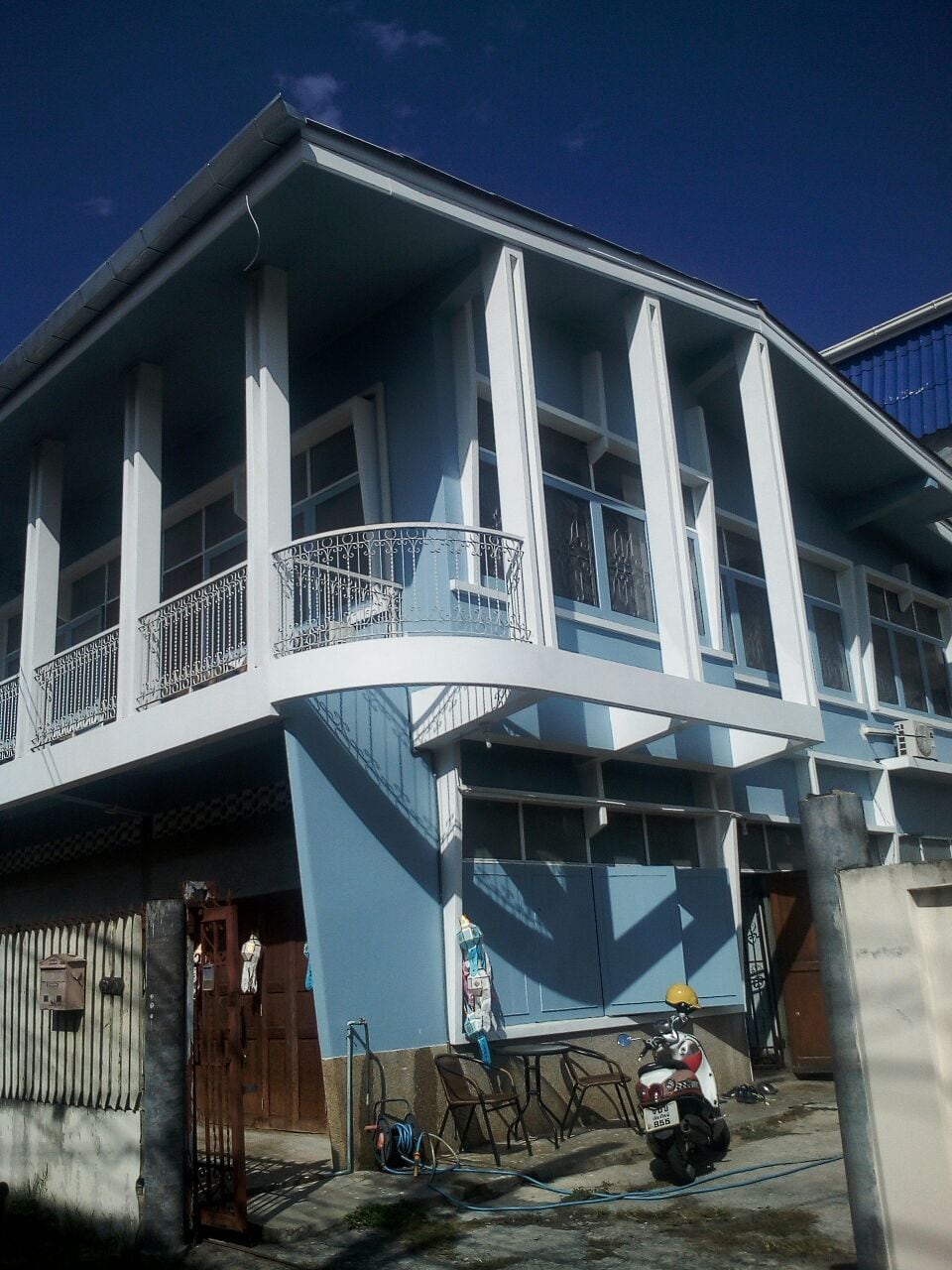 The exterior of a light blue house features distinctive architectural elements, including a balcony with decorative railings. Multiple large windows allow natural light to fill the space. The surrounding area includes a motorcycle parked at the entrance and chairs positioned outside for relaxation.
