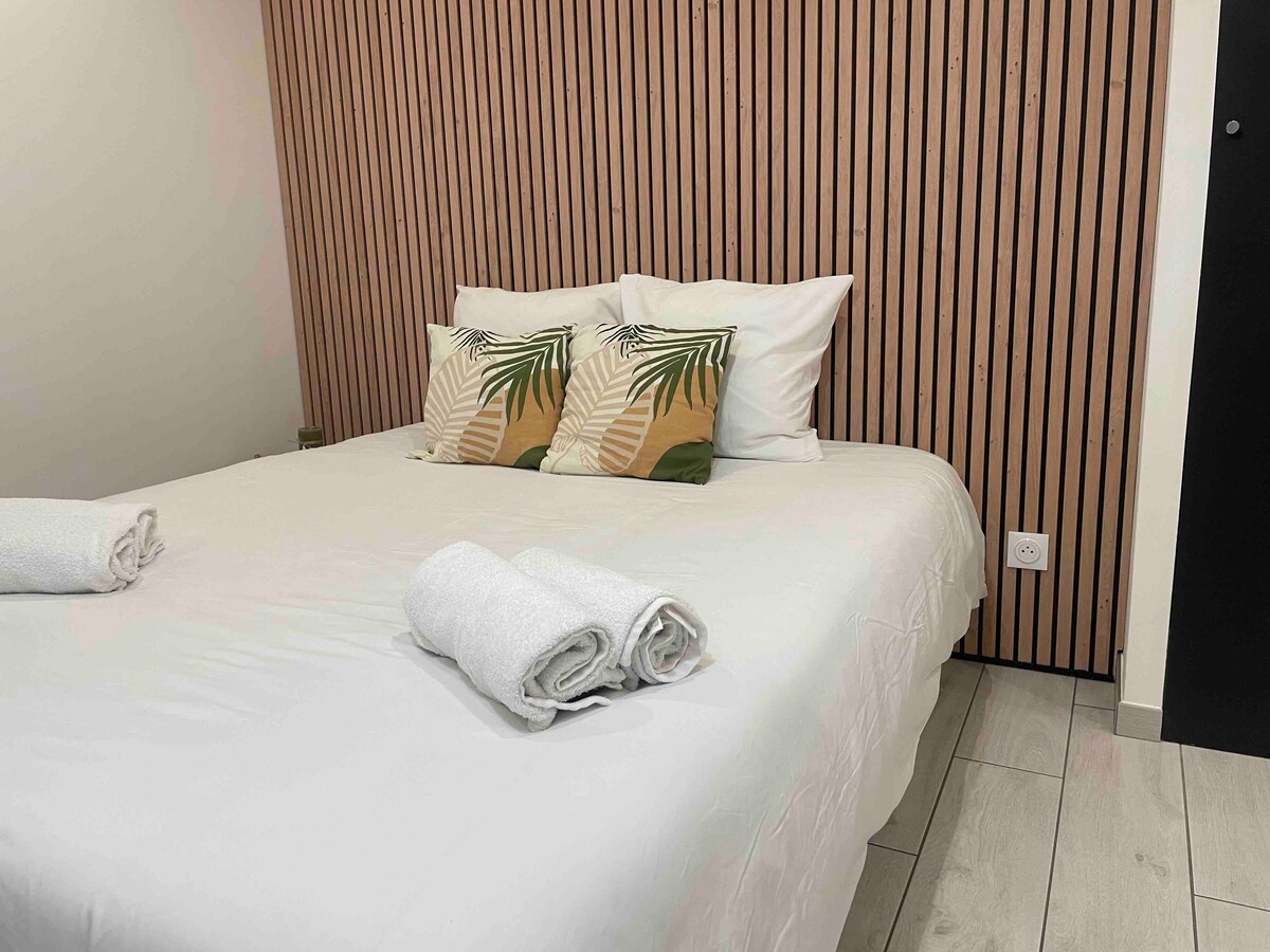 A spacious bed is dressed with crisp linens, flanked by two decorative pillows featuring tropical leaf patterns. Soft, rolled towels are neatly arranged at the foot of the bed. The room's walls are adorned with vertical wooden slats, contributing to a modern and fresh aesthetic.
