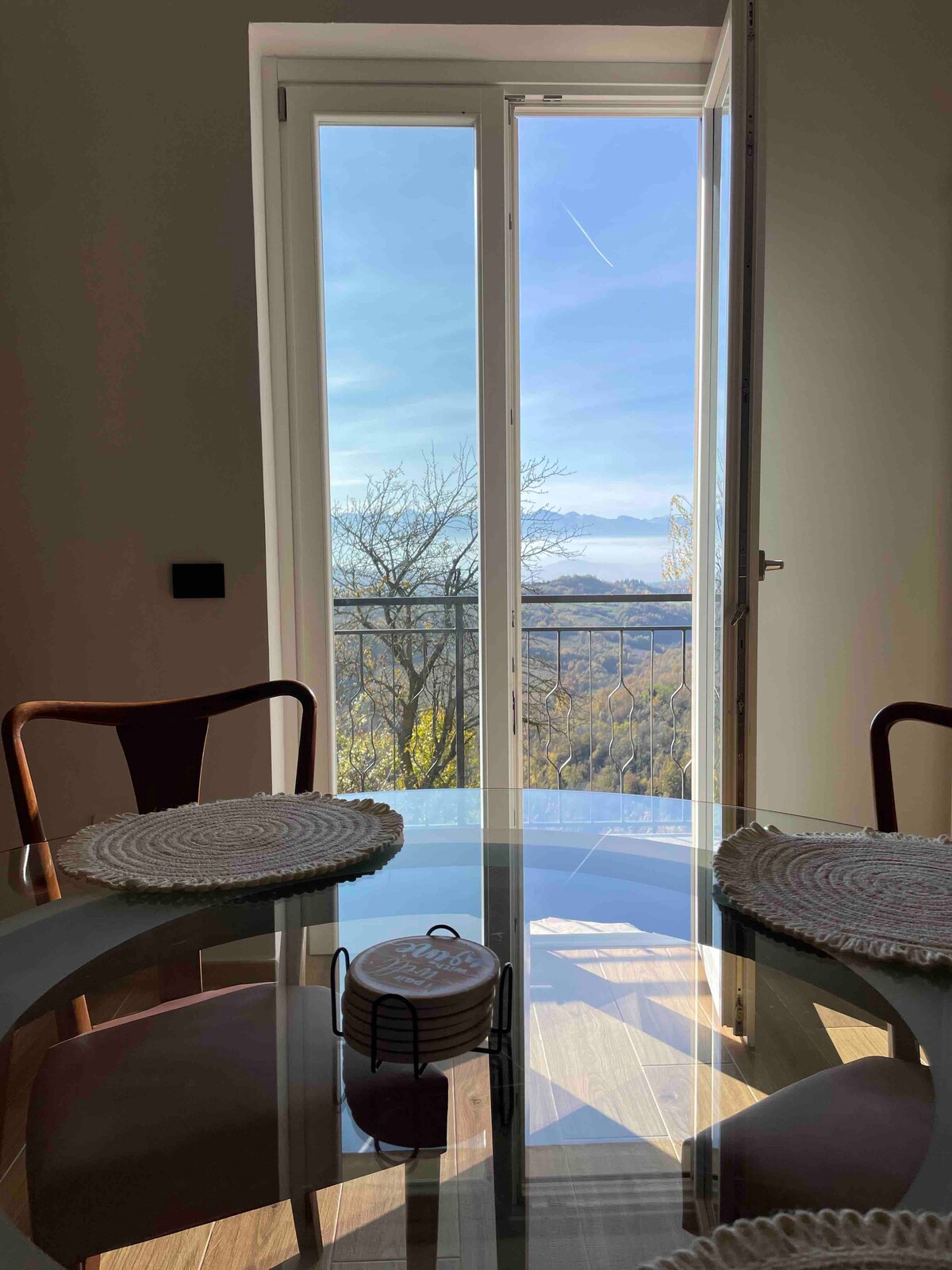 A dining area is presented with a round glass table surrounded by wooden chairs. Natural light enters through a glass door that opens to a balcony, providing a view of the landscape beyond. A decorative item sits at the center of the table.