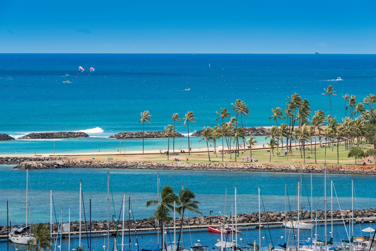 A panoramic view showcases the tranquil Pacific Ocean under a clear blue sky. In the foreground, a marina filled with sailboats is visible, while the sandy beach and grassy areas lined with palm trees create a serene coastal landscape. Brightly colored parachutes can be seen in the distance.