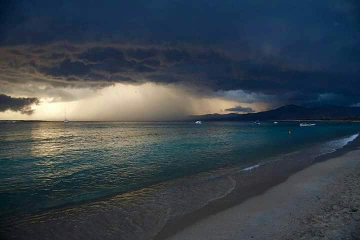 A panoramic view of the coastline is presented, with dark clouds looming overhead. The water displays various shades of blue and turquoise, reflecting the sky. A sandy shore lines the beach, while distant boats dot the water, creating a sense of depth in the landscape.