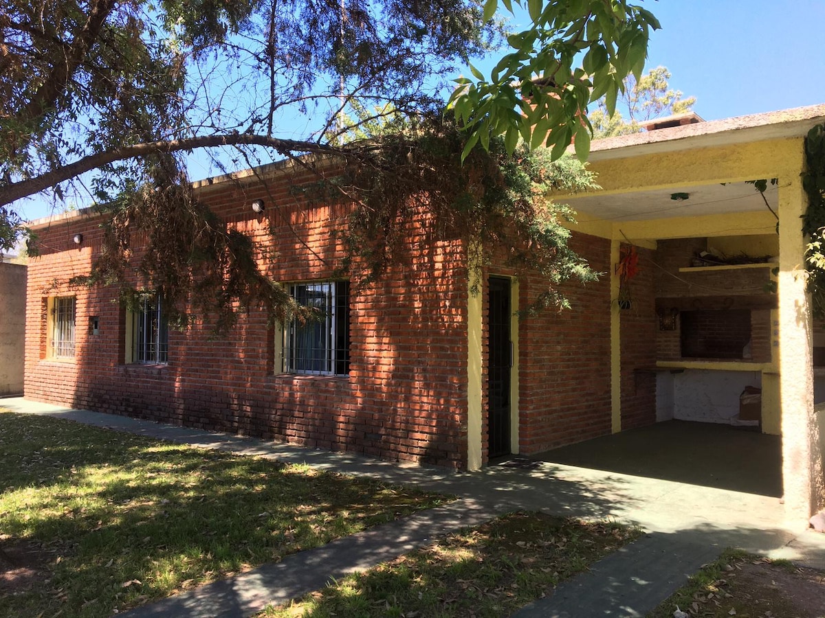 The exterior of the home is constructed of brick, featuring large windows and a covered patio area. A barbecue area is integrated into the structure. Lush grass is visible, complemented by the shade of surrounding trees.