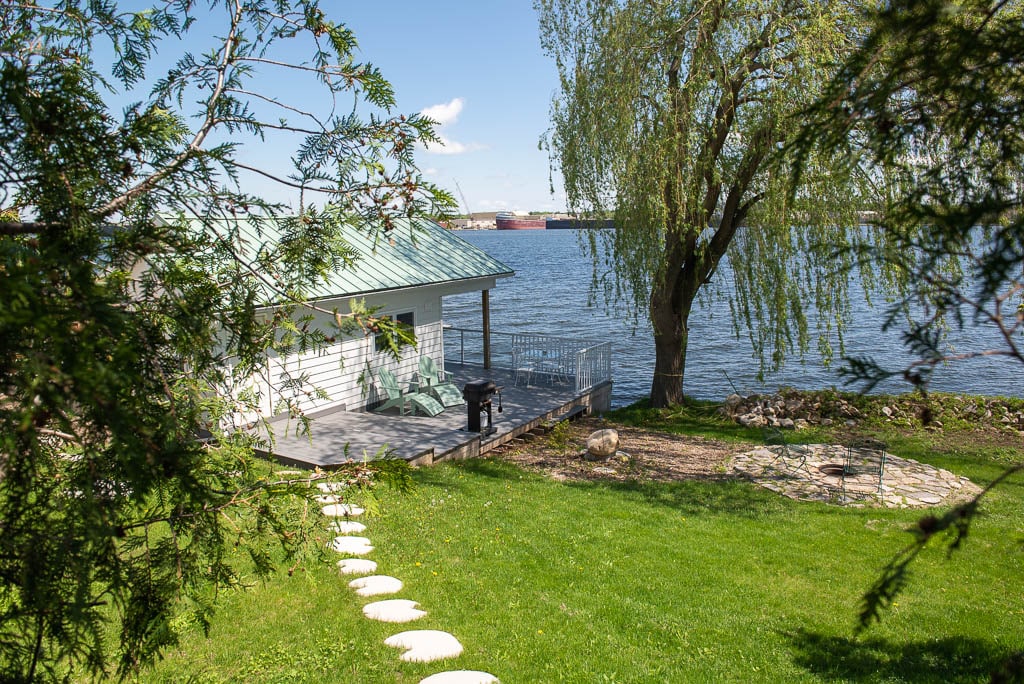 The image shows a serene waterfront scene featuring a cottage with a green metal roof. A spacious deck is equipped with a grill, complemented by a circular stone fire pit in the yard. Lush greenery surrounds the area, gently leading down to the water's edge.