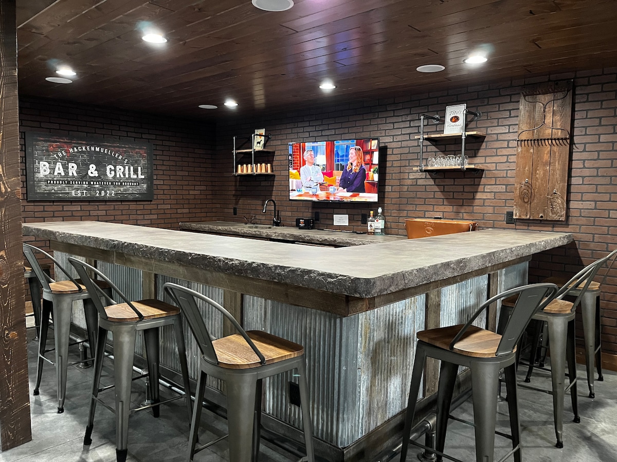 A modern home bar area is featured with a large granite countertop surrounded by multiple metal stools. A flat-screen television is mounted on the wall, and rustic decor elements, including a vintage bar sign and wooden accents, add character to the space. Warm lighting enhances the inviting atmosphere.