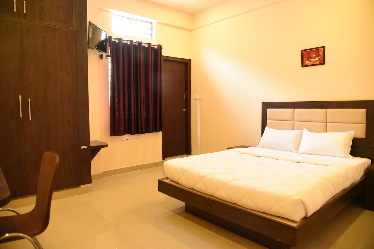 A spacious sleeping area features a large bed with a white quilted cover. Dark curtains frame a window, while a mounted television is visible. A simple wood desk and chair complement the room's modern design.