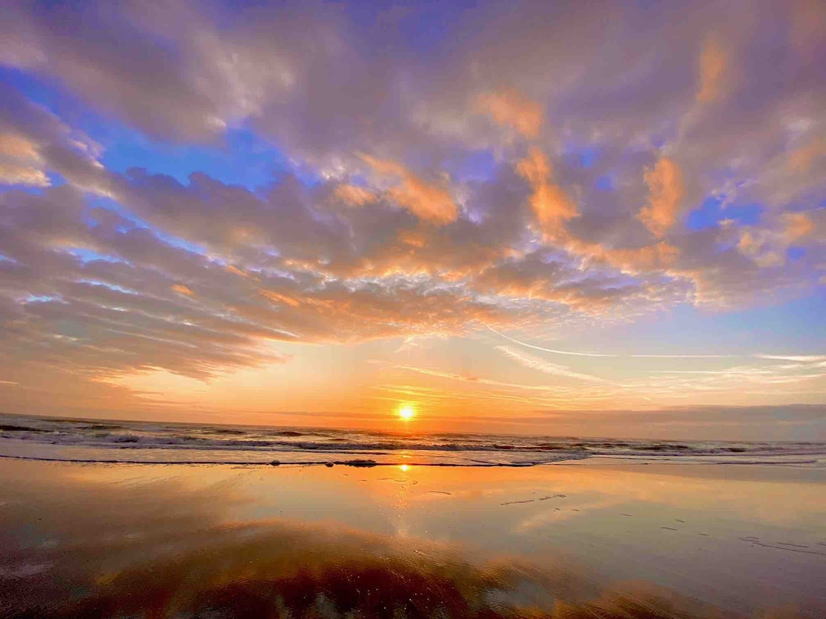 A beautiful sunset is captured over the ocean, showing vibrant hues of orange and pink reflecting on the wet sand. Soft clouds are scattered across the sky, creating a calm and serene atmosphere as the waves gently lap at the shore.