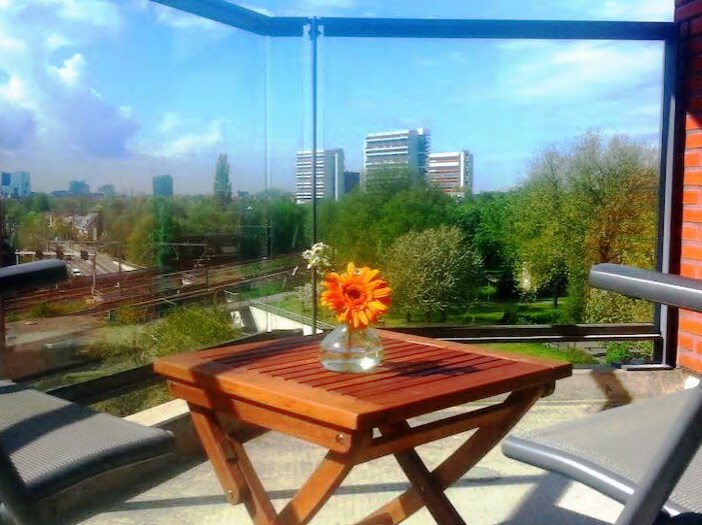 A private terrace is featured, showcasing a small wooden table with a vase holding a sunflower. Comfortable chairs are positioned around the table, and expansive glass panels provide a view of green trees and distant buildings under a bright sky.