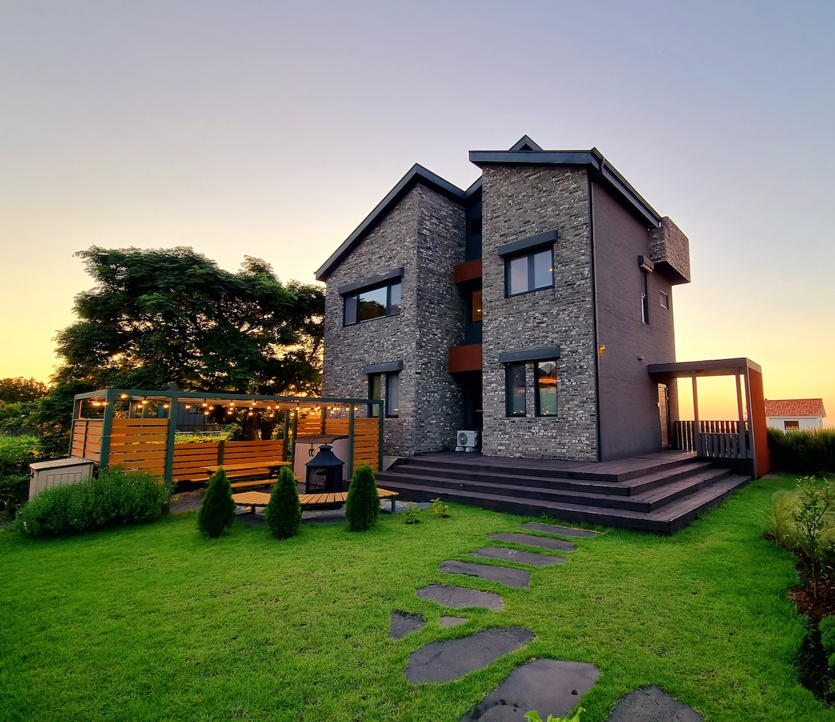 A modern two-story house is framed by lush greenery, featuring a combination of stone and dark exterior walls. A spacious deck and patio area provide outdoor seating options, while well-maintained landscaping enhances the overall appeal of the property.