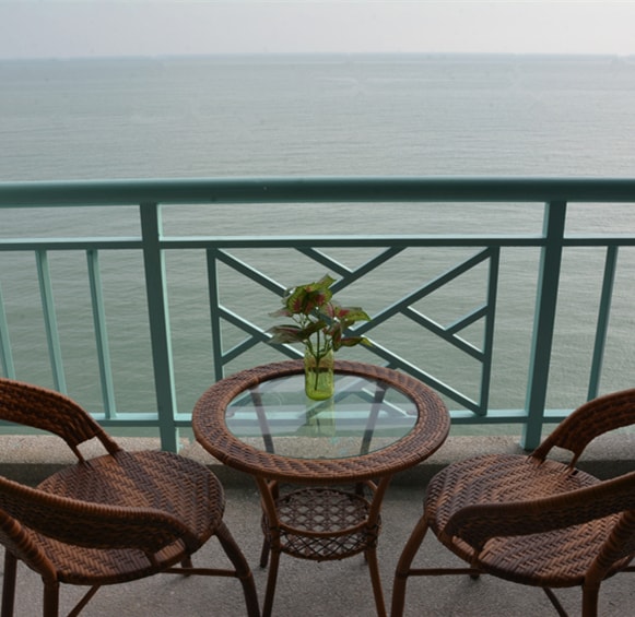 The balcony showcases a cozy seating arrangement with two woven chairs and a round glass table. A small plant in a glass vase sits atop the table, with a panoramic view of the sea extending towards the horizon.