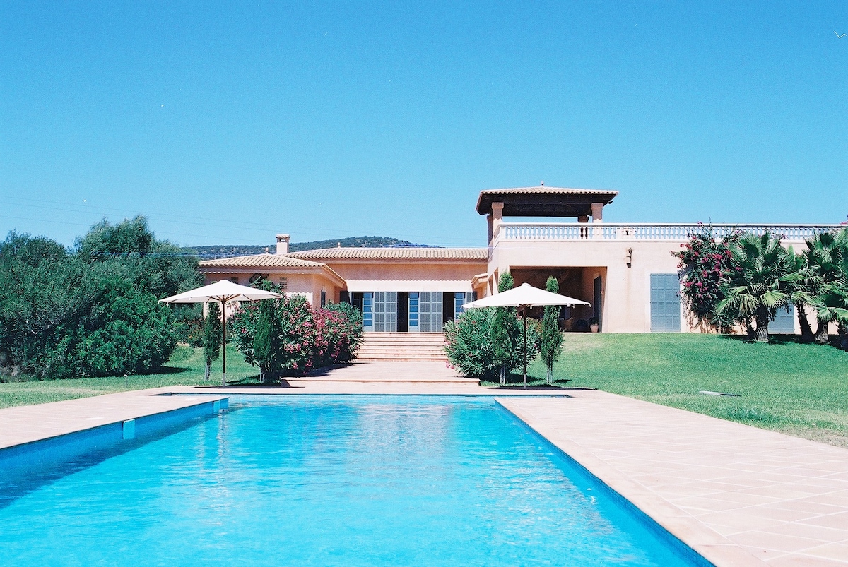 A large swimming pool stretches across the foreground, reflecting clear blue skies. Surrounding greenery includes well-maintained bushes and palm trees, creating a tranquil outdoor setting. The estate's main building features a spacious porch and multiple entrances, harmoniously integrated into the landscape.