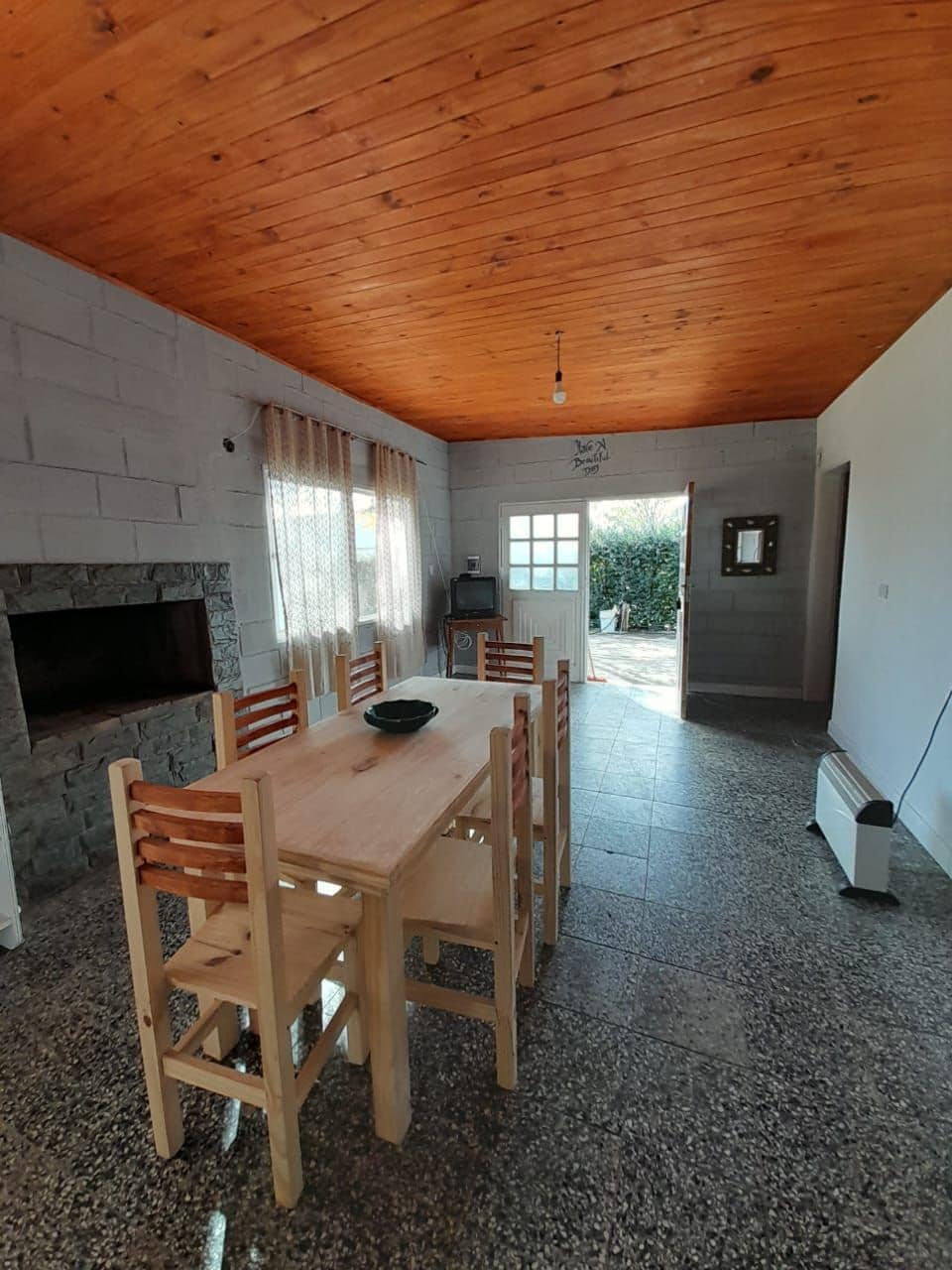 A dining area features a wooden table surrounded by six chairs, with a stone fireplace on one side. Large windows allow natural light to fill the space, and a door leads to a garden area outside. The ceiling showcases warm wooden beams.