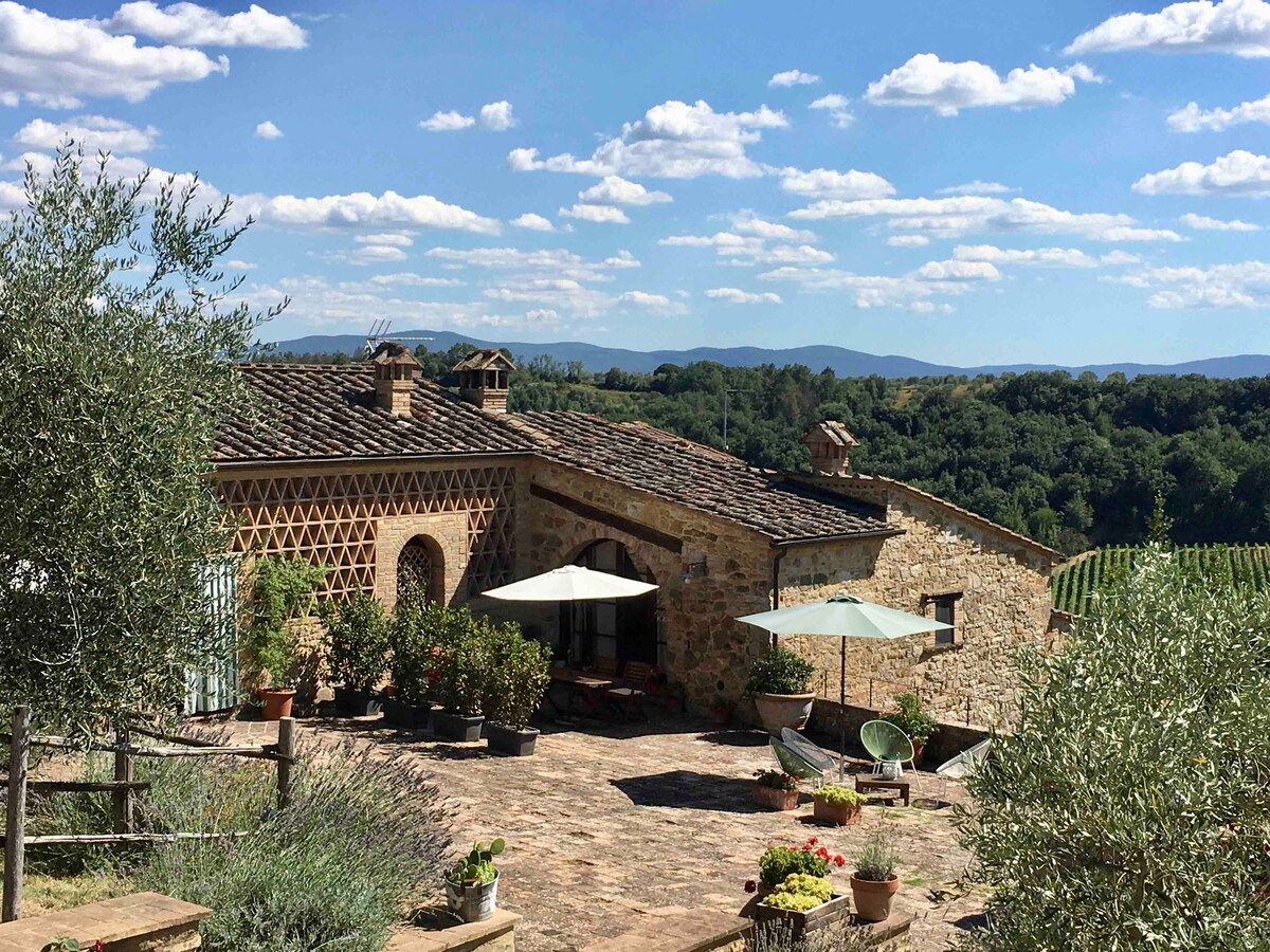 A rustic stone building is seen surrounded by greenery, with patio areas featuring umbrellas and potted plants. The landscape includes vineyards and rolling hills under a blue sky with scattered clouds, creating a serene outdoor space perfect for relaxation.