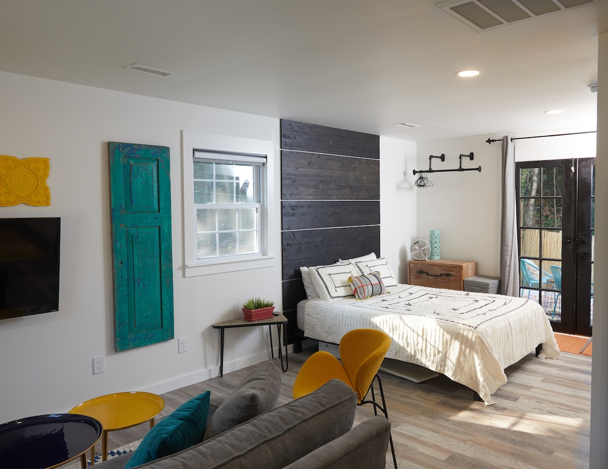 The spacious suite features a queen bed with a patterned bedspread, complemented by a cozy love seat and two accent chairs. A flat-screen TV is mounted on the wall, and decorative elements include a colorful door and a wooden accent wall. Natural light enters through the window and sliding door.