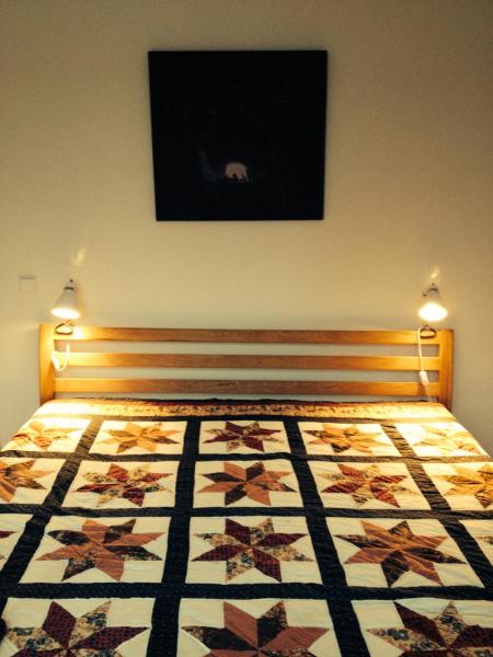 A wooden bed frame supports a quilt featuring a star pattern in warm colors. Two bedside lights provide soft illumination, and a dark, abstract artwork is hung above the headboard. The surrounding walls are painted in neutral tones, contributing to a calming environment.