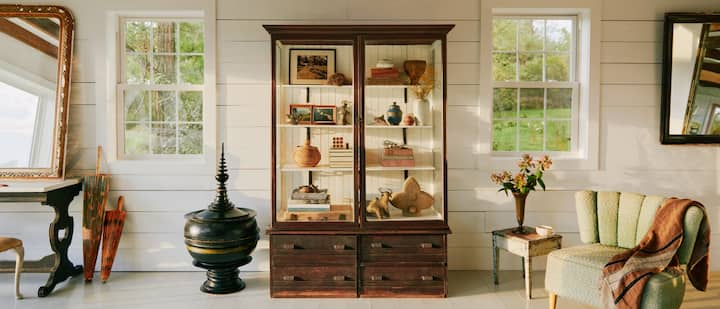 A photo shows a large white living room with a collection of antique furniture, including a large wood and glass display case full of an assortment of vintage valuables.