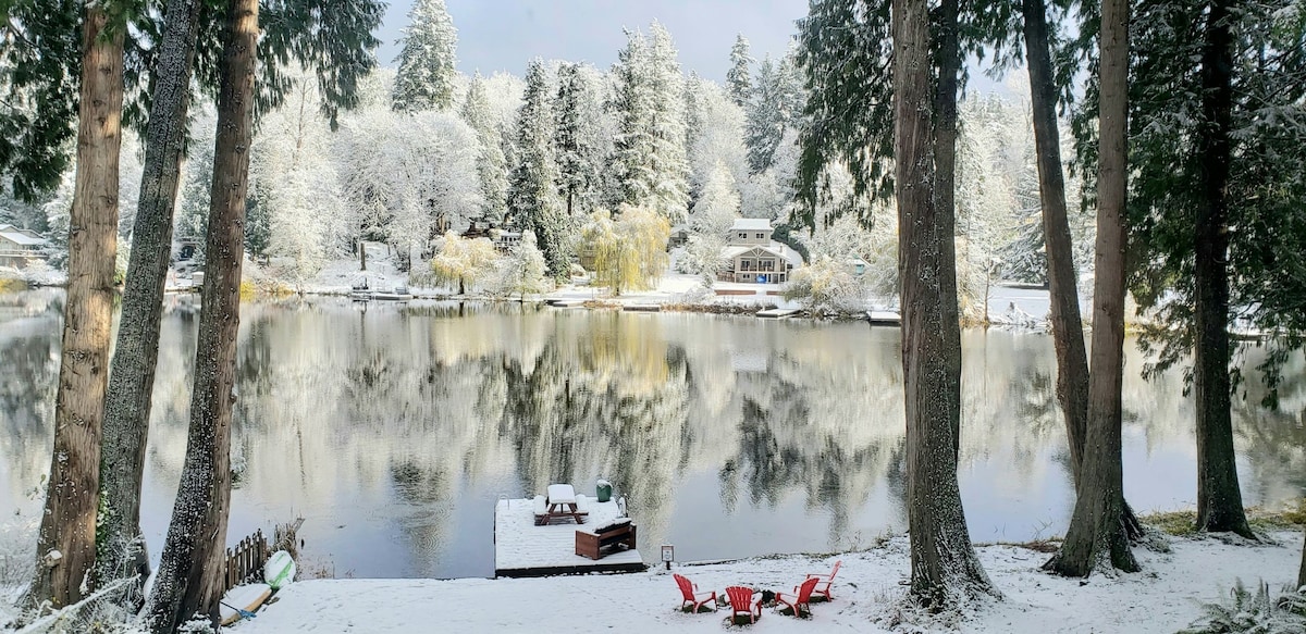 A serene lakeside view is captured, featuring a snowy landscape. Tall pine trees frame the image, with a still lake reflecting the surrounding scenery. A wooden dock is visible in the foreground, accompanied by colorful chairs. Snow blankets the ground and the trees.