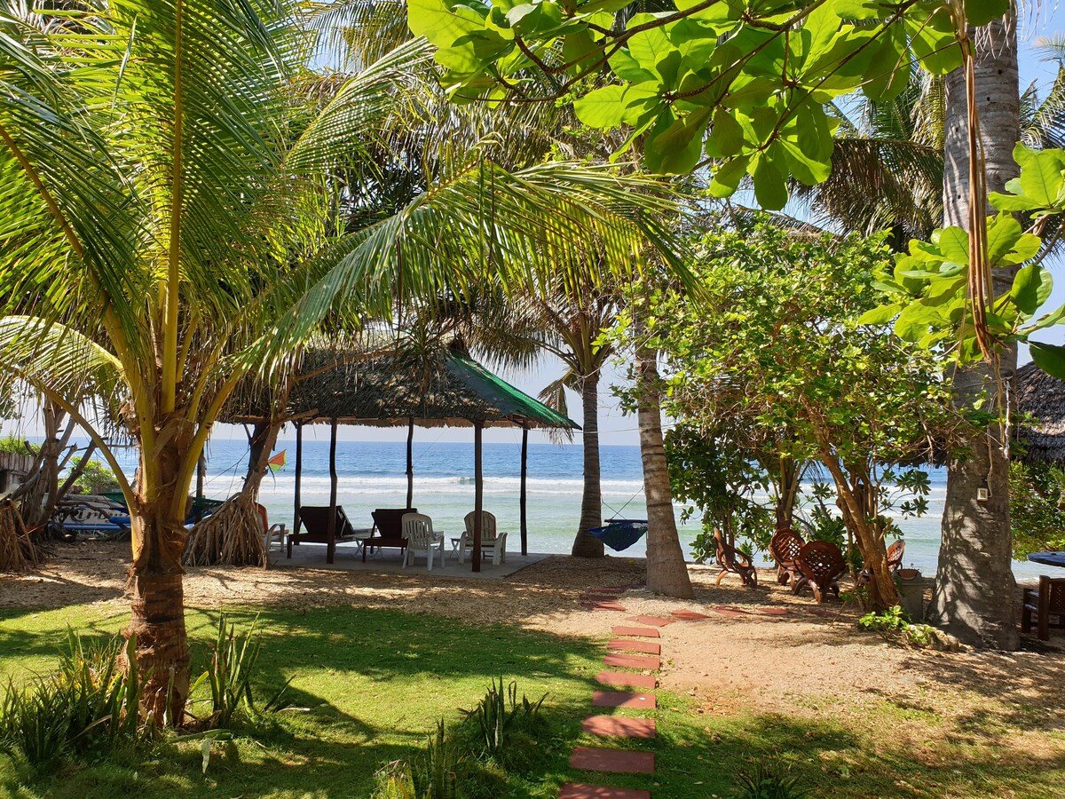 A serene garden area is framed by lush palm trees and tropical plants. A thatched-roof gazebo is visible in the background, providing seating for relaxation. Soft sands and gentle waves can be seen beyond, creating a tranquil coastal atmosphere.