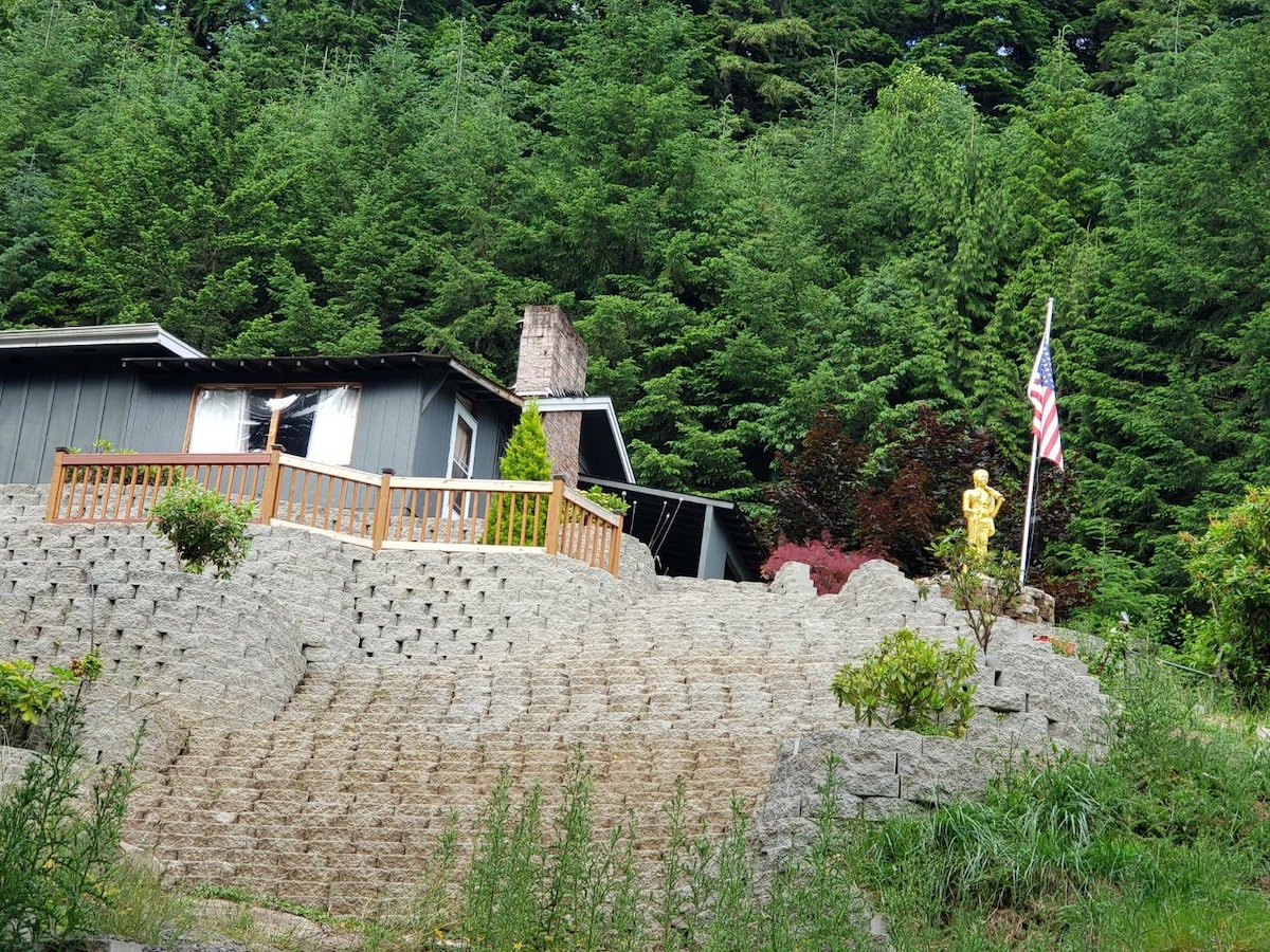 A hillside property is surrounded by lush greenery, featuring a stone pathway that leads to a spacious wooden deck. A golden statue and American flag are notable accents on the deck, with a backdrop of tall trees enhancing the serene environment.