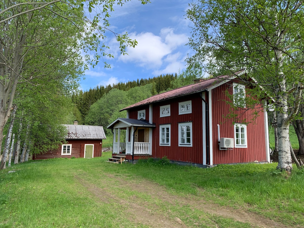 A charming red house is positioned amidst greenery, featuring multiple windows and a front porch. A smaller structure is visible nearby, under a clear blue sky dotted with clouds. The surrounding landscape includes trees and a grassy area, enhancing the serene environment.