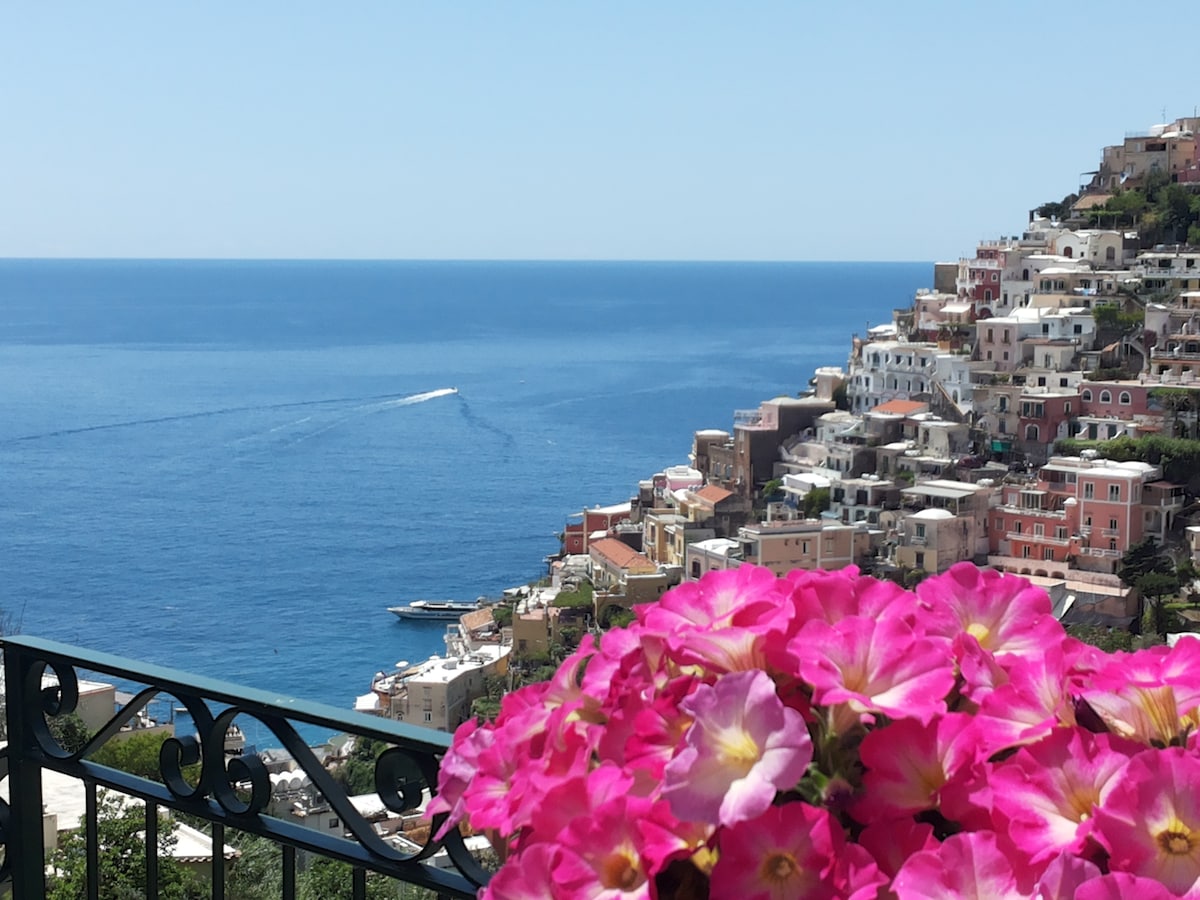 A vibrant cluster of pink flowers frames a panoramic view of the blue ocean and the coastal town of Positano. Colorful buildings cascade down the hillside, with boats visible on the water's surface. A clear sky enhances the serene atmosphere of the scene.