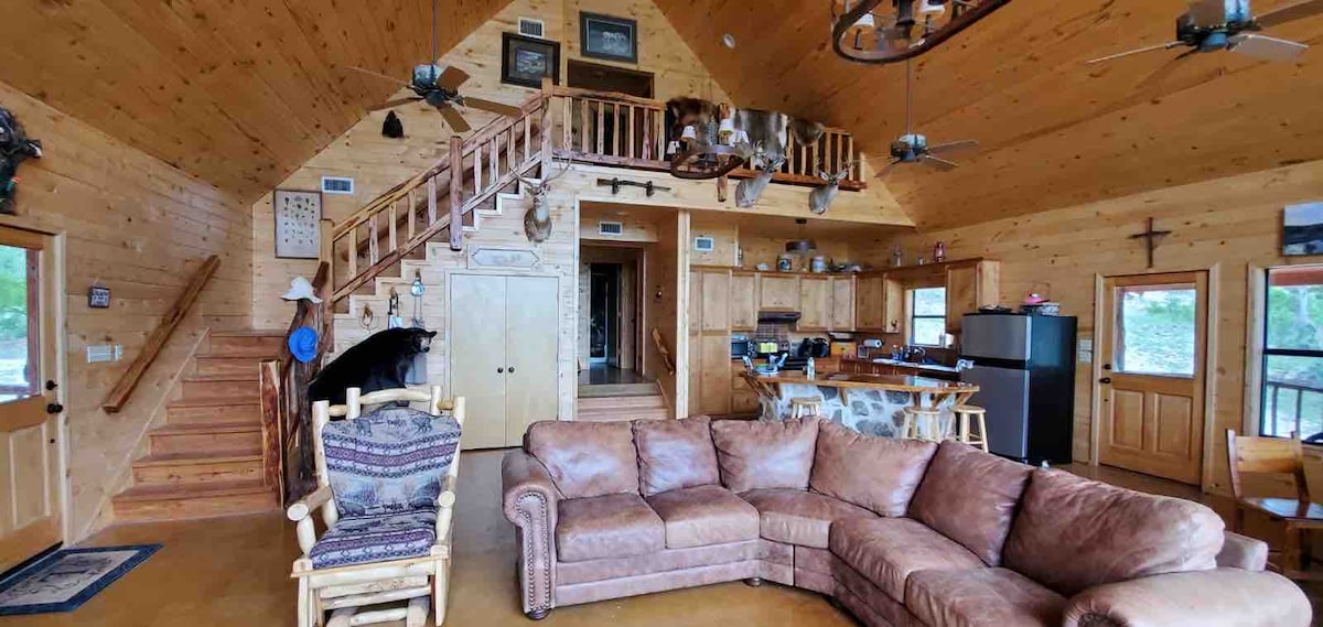 A spacious living area features a large sectional sofa arranged around a coffee table, complemented by wooden walls and exposed beams. A staircase leads to an upper level, and a view of the kitchen and dining area is visible in the background.