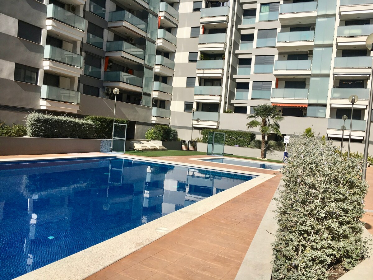 A clear view of the residential pool area is presented, with two swimming pools bordered by sunlit tile pathways. The surrounding greenery provides a serene backdrop, and modern apartment buildings are visible in the background, contributing to the calm atmosphere of the space.