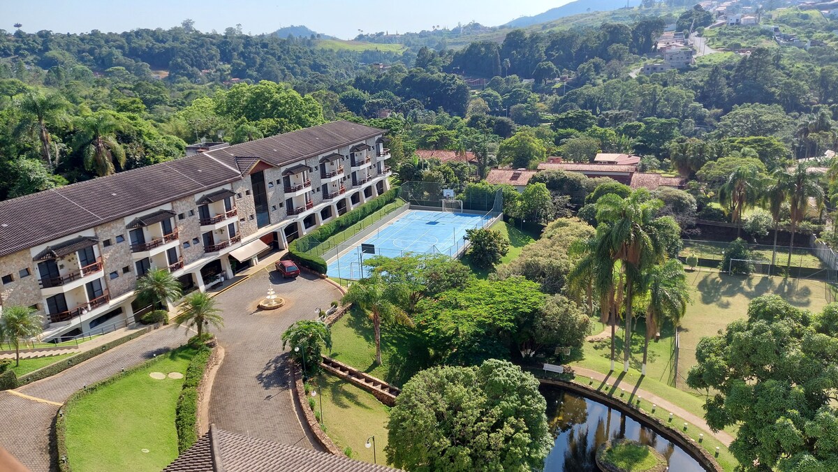An aerial view captures the landscaped grounds, featuring lush greenery and a swimming pool surrounded by tennis courts. The multi-story building showcases natural materials, complemented by paths leading through the grounds, creating an inviting outdoor environment.