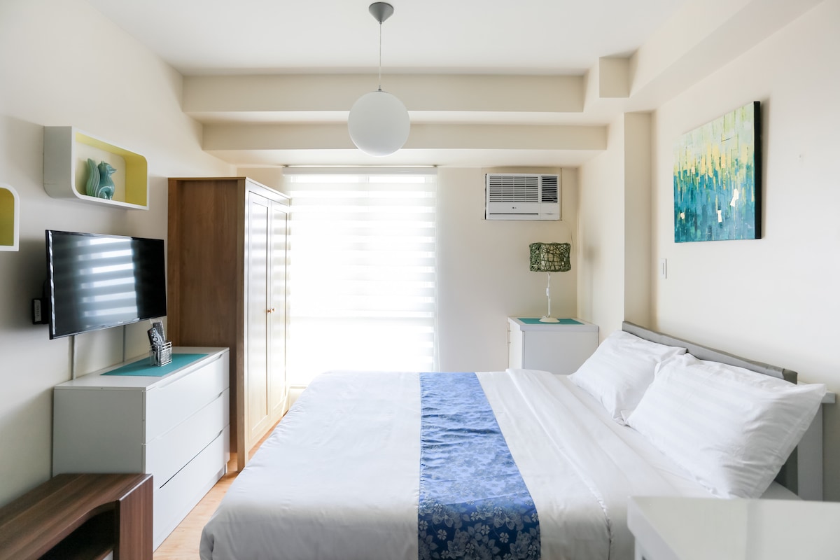 The bedroom features a spacious queen-sized bed adorned with crisp white linens and a blue decorative runner. A flat-screen TV is mounted on the wall across from the bed. A large closet and dresser provide ample storage, while natural light filters through sheer window shades.