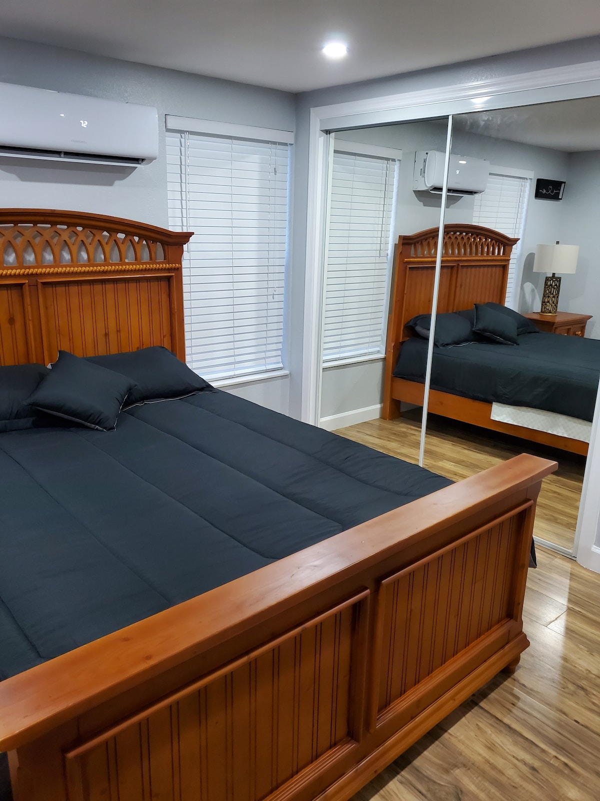 A cozy sleeping area features a wooden bed frame with a dark bedspread and two pillows on a spacious bed. The room contains a mirrored closet reflecting the interior and is illuminated by soft overhead lights. Large windows with blinds provide natural light.