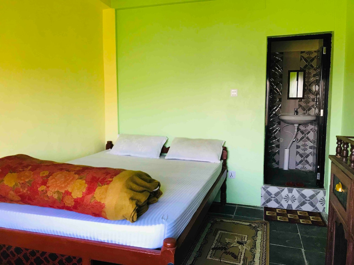A spacious bedroom is presented with vibrant green walls, featuring a comfortable bed with soft pillows. A patterned blanket drapes over the bed. An adjoining private bathroom is accessible through a door, visible in the corner of the image.