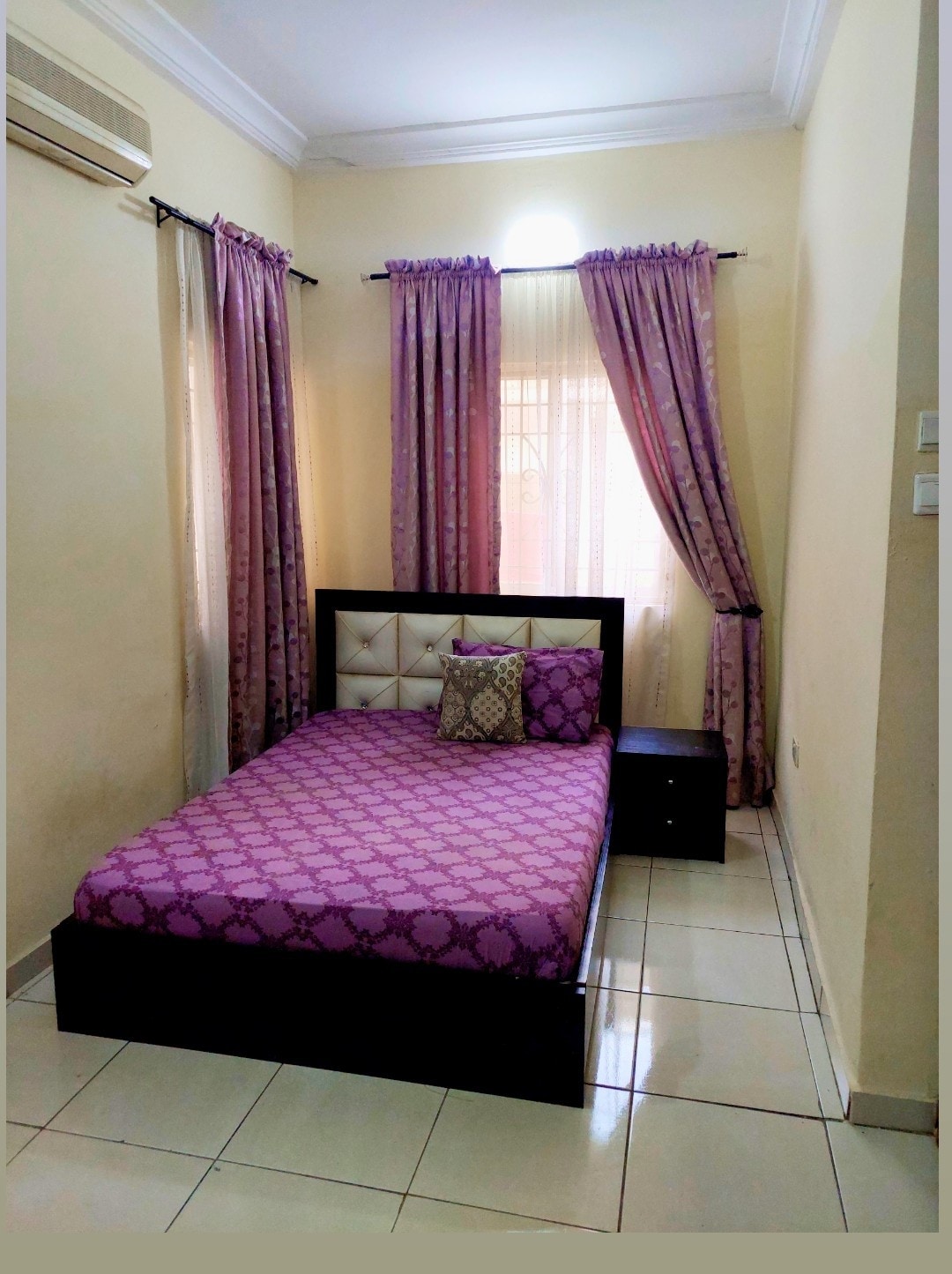 A cozy sleeping area features a spacious bed with a purple patterned coverlet. A decorative cushion adds a touch of style. Natural light filters through sheer curtains, illuminating the room. A bedside table is positioned next to the bed, with walls finished in a soft beige tone.