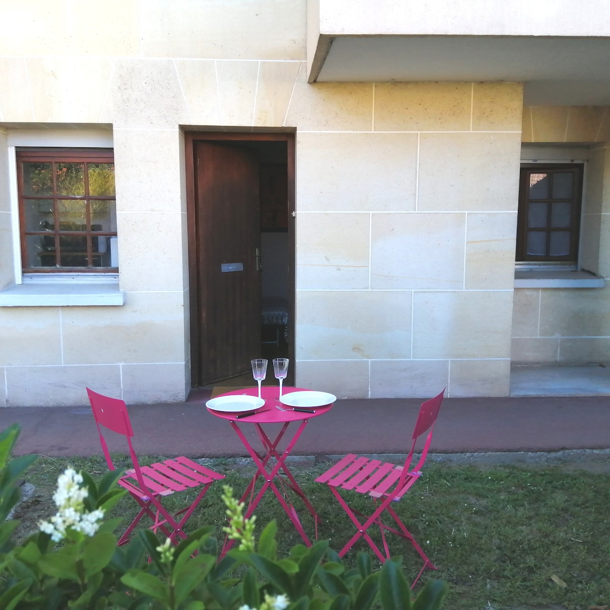 A small patio area features a round table with two pink folding chairs. Two plates and two champagne flutes are placed on the table. The independent entrance to the studio is visible, flanked by two windows, amidst a small patch of lawn.