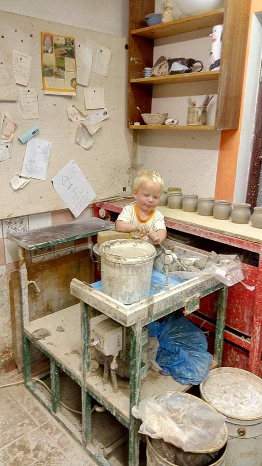 A pottery workspace is visible, featuring a potter's wheel at the center surrounded by various clay containers and tools. A notice board is mounted on the wall, displaying papers and notes. Shelves above hold organized pottery items, creating a creative environment.
