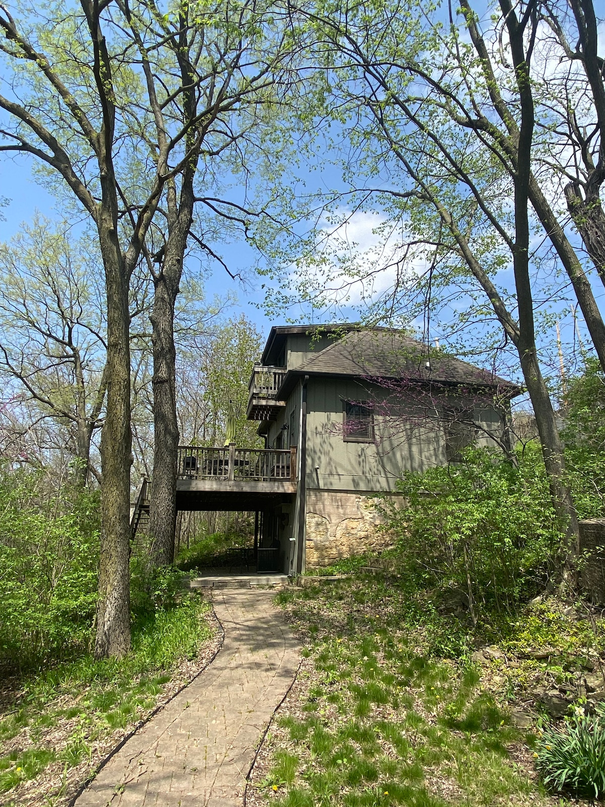 A secluded hilltop house is surrounded by trees and greenery. A pathway leads to the two-story structure, which features a small balcony on the second floor. Natural light filters through the leaves, creating a serene outdoor atmosphere.