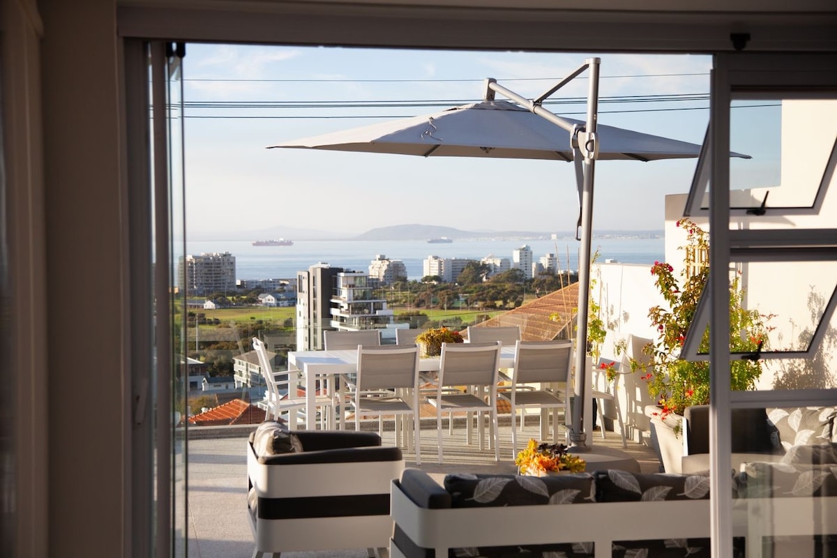 An expansive outdoor terrace is featured, showcasing a dining area with a large table and chairs under a sleek umbrella. The area overlooks a scenic view of the ocean and surrounding city, with greenery and flowers adding natural beauty to the setting.