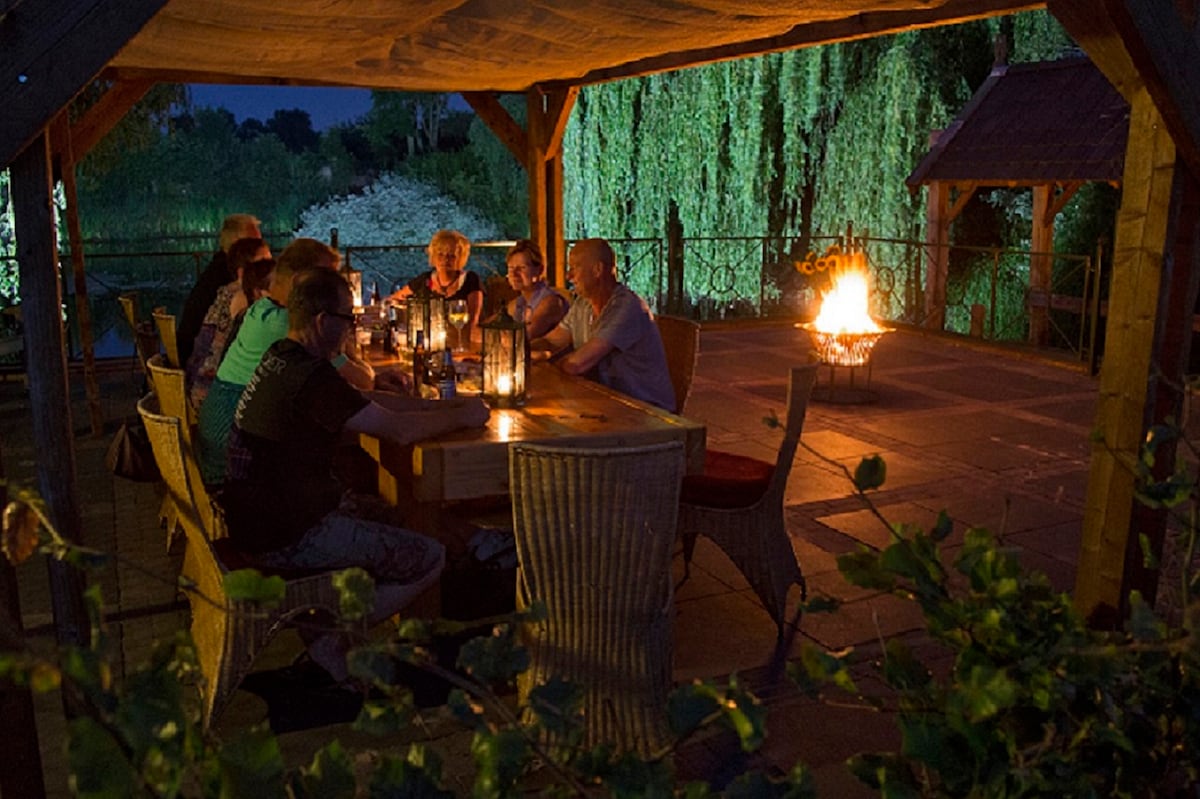 An outdoor dining area is presented under a wooden pergola, illuminated by lanterns and a warm fire. A large table, surrounded by chairs, is occupied by guests enjoying dinner, with lush greenery visible in the background, creating a serene evening atmosphere.