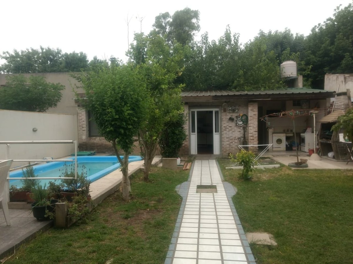 A spacious outdoor area features a well-maintained swimming pool surrounded by greenery. A path of pavers leads to the entrance of a cozy cabin-style loft. Nearby, a shaded seating area is visible, offering a relaxing space beneath the trees.