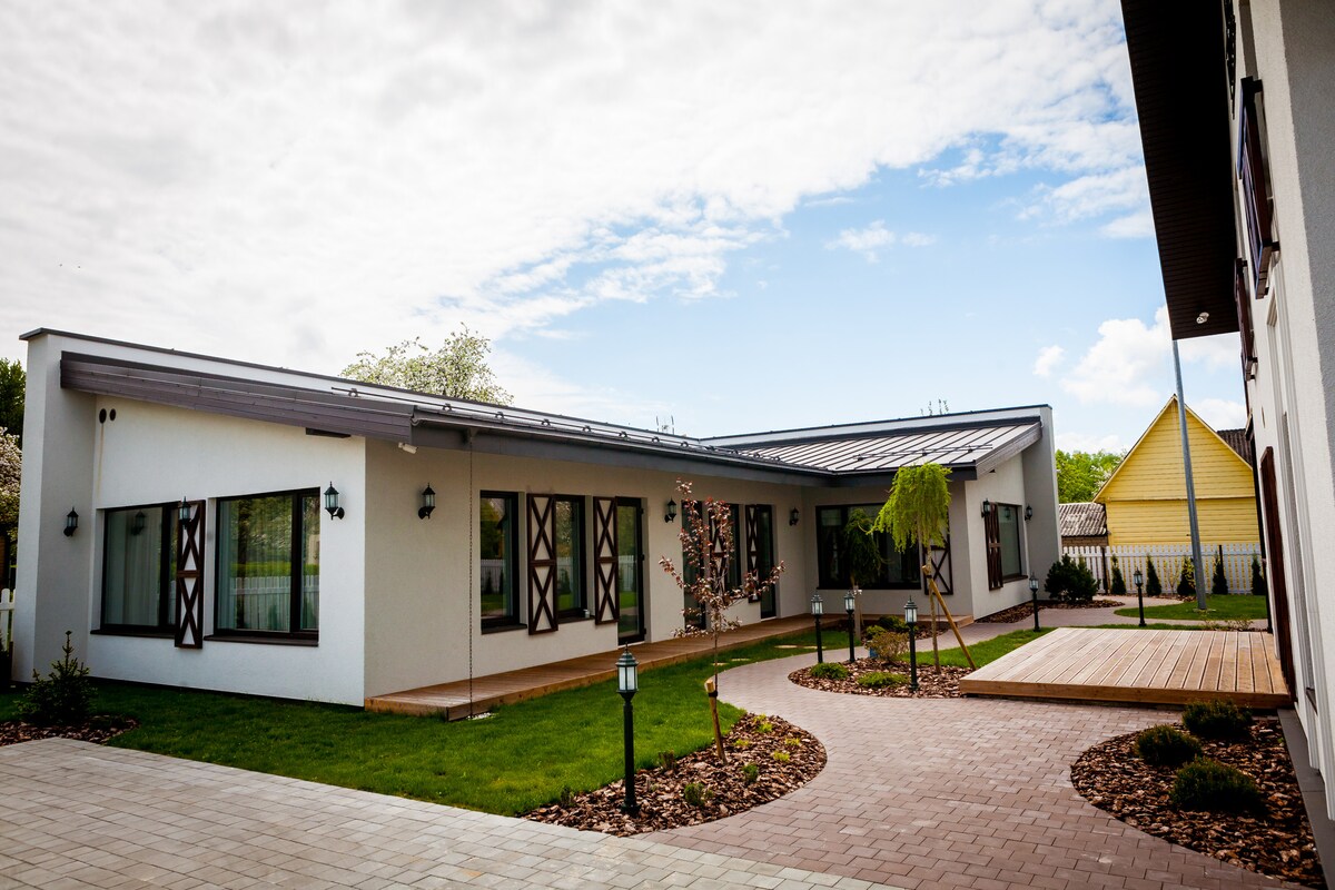 A modern villa is presented with a landscaped courtyard featuring a paved path winding through green grass and decorative shrubs. Large windows are set into light-colored walls, reflecting the outdoor light, while a wooden deck extends from the building, creating an inviting outdoor space.