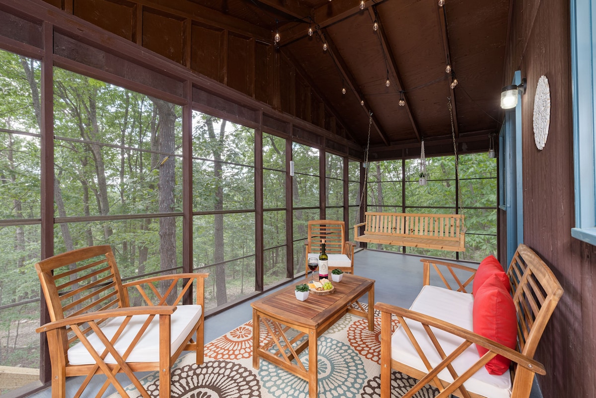 A screened porch is presented with wooden seating and a welcoming swing. Natural light filters through the screens, illuminating the space. A round area rug adds a touch of color, with a small table set for refreshments, surrounded by views of the green forest.