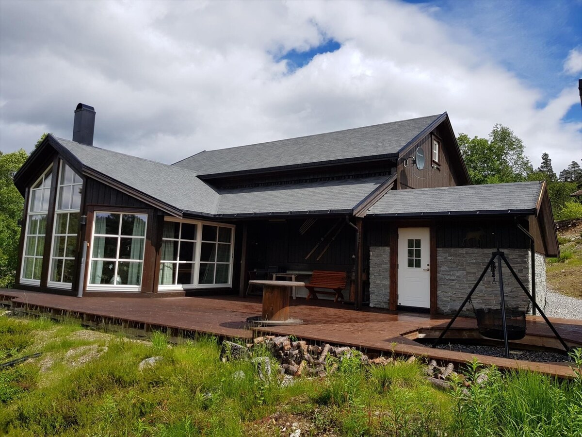 The exterior of the cabin features a spacious wooden deck, complemented by large windows that provide ample natural light. A swing set is visible on the deck, and a stone wall accentuates the entrance. The surrounding landscape is lush with greenery.