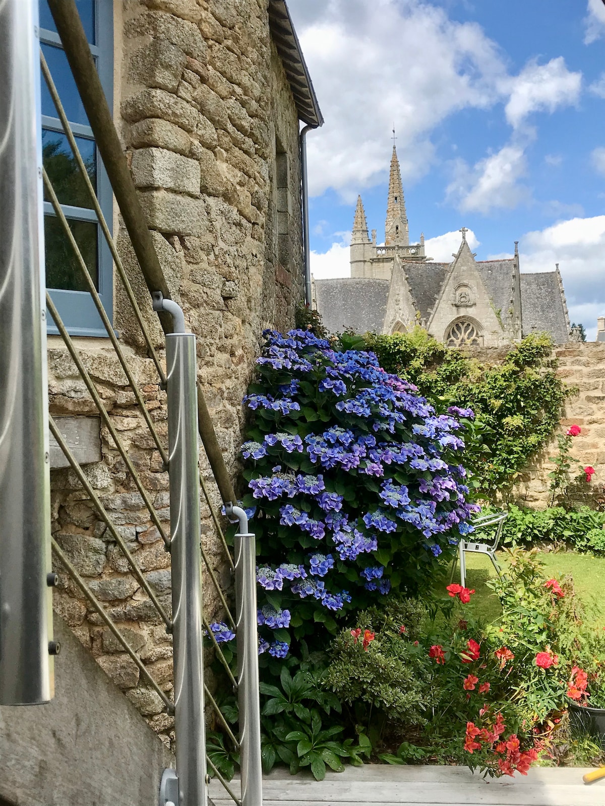 A stone exterior is shown, featuring a stainless steel handrail leading down to a garden area. Vibrant blue hydrangeas bloom alongside red roses, bordered by lush greenery. In the background, the spires of a church rise against a partly cloudy sky.