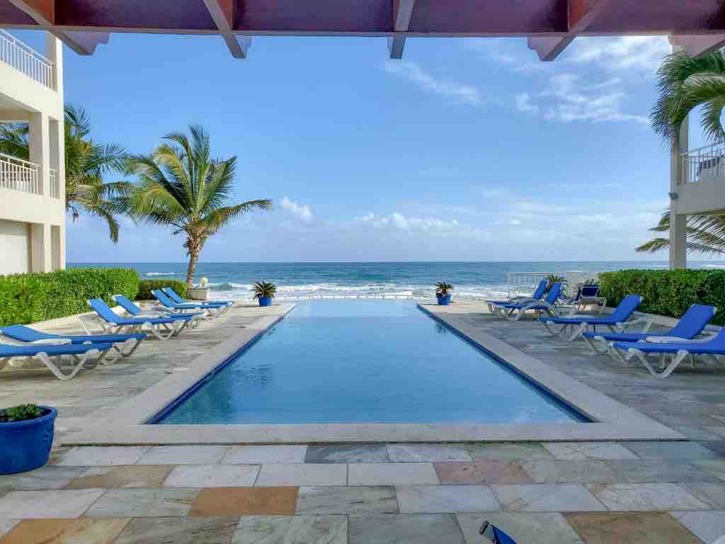 An infinity pool stretches towards the ocean, framed by palm trees. Lounge chairs line the pool area, inviting relaxation. The expansive sky reflects in the calm water, enhancing the serene beach view beyond. A well-maintained border of greenery surrounds the space.