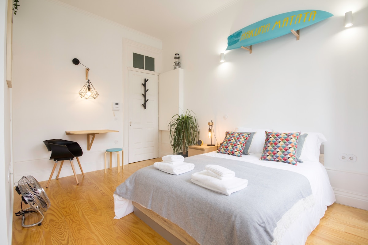 A spacious studio is highlighted by a comfortable bed adorned with decorative pillows. A small dining table and a chair are situated against the wall, complemented by decorative lighting. A surfboard is mounted above the bed, adding a unique touch to the minimalist decor.